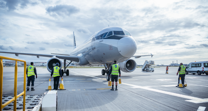 Four ground crew members in reflective vests guide a commercial airplane as it taxis on the airport tarmac under a cloudy sky.