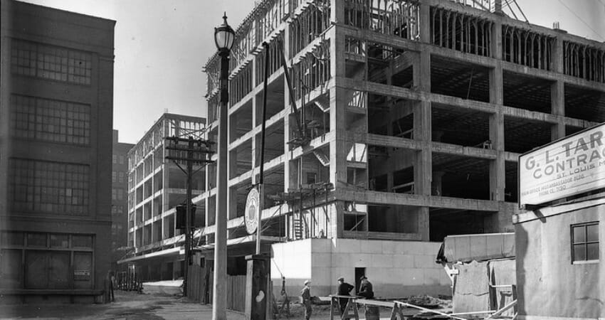 Black and white photo of a multi-story building under construction, surrounded by scaffolding, workers, and a sign for a construction contractor.