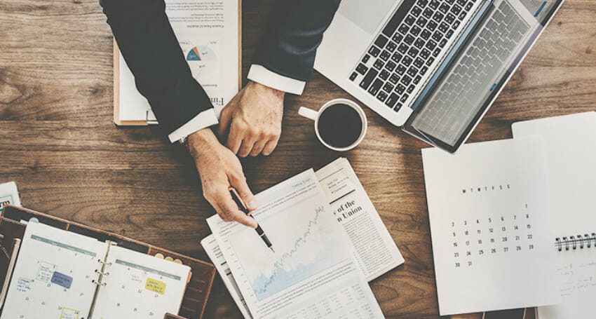 A person in business attire reviews graphs and documents at a desk with a laptop, calendar, coffee cup, and open planners.