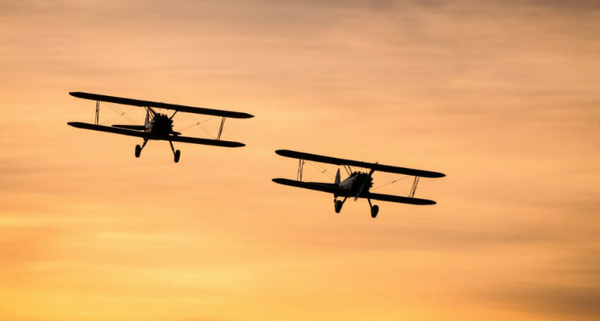 Two vintage biplanes are flying side by side against an orange sky at sunset.